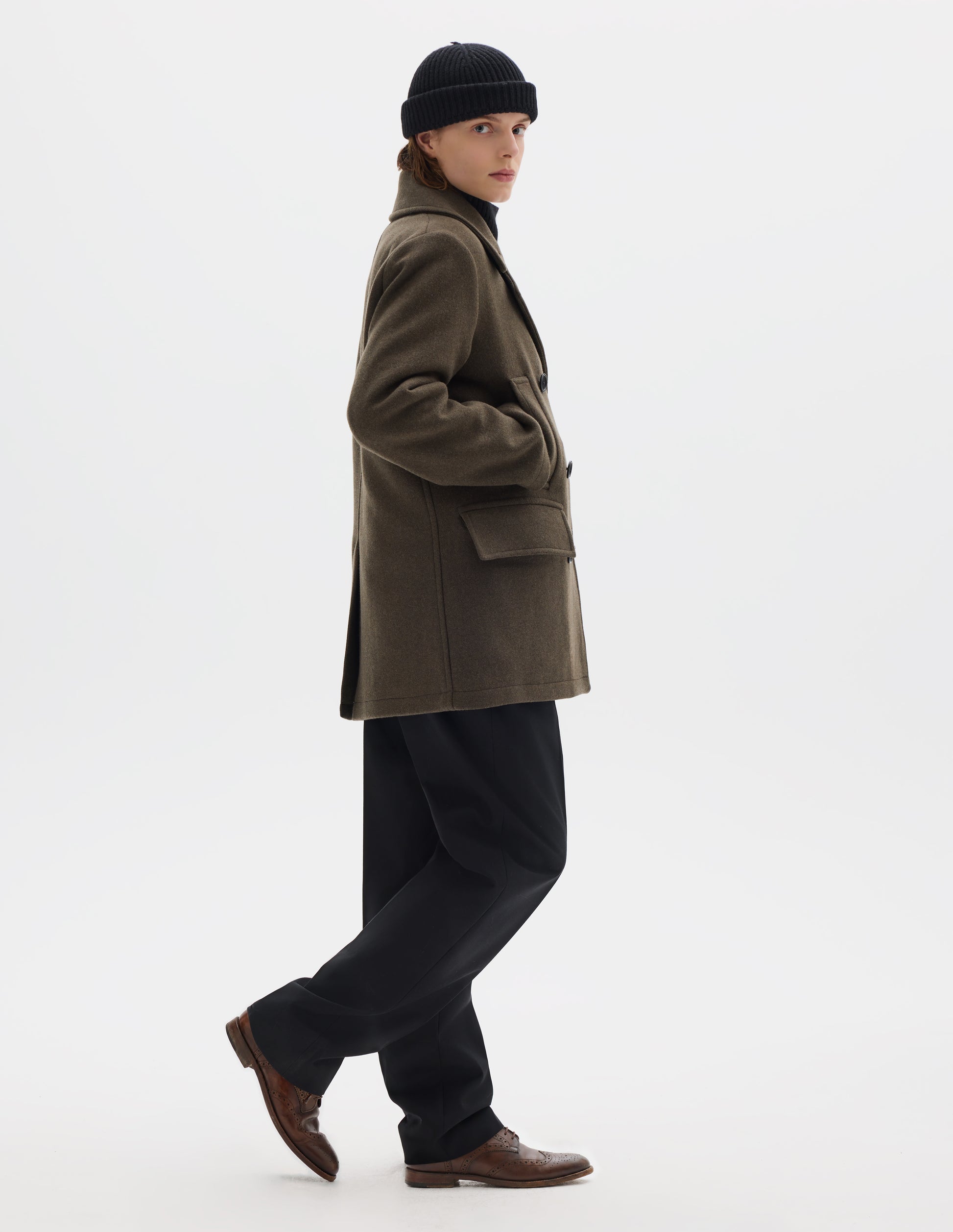 Full body image, side view of model wearing khaki green wool blend coat with large collar and revere, horn buttons, high-set single welt pockets and large flap pockets, and with rear vent. Styled with black beanie, black trousers and brown leather brogues.