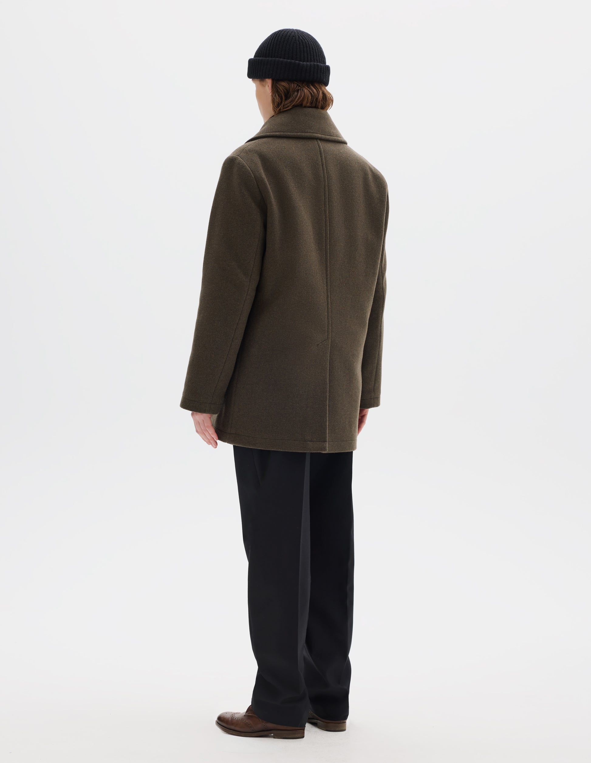 Full body image, back view of model wearing khaki green wool blend coat with large collar, centre back seam and rear vent. Styled with black beanie, black trousers and brown leather brogues.