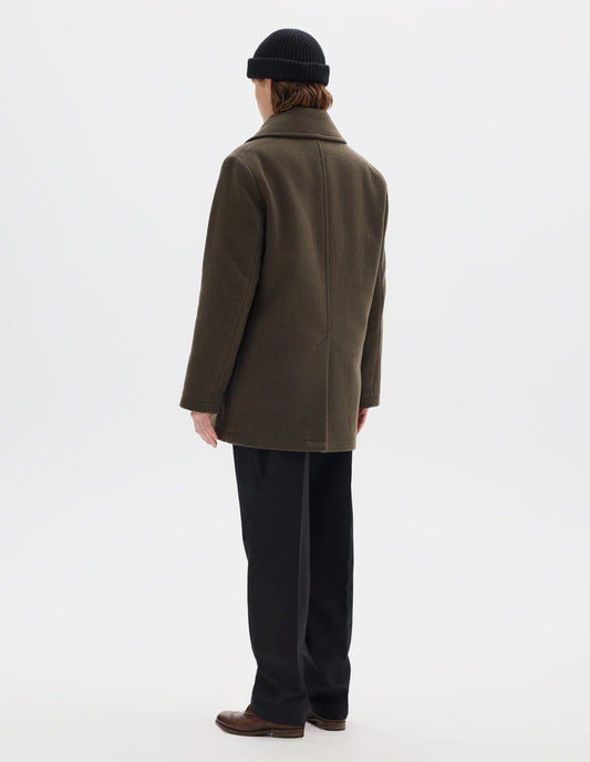 Full body image, back view of model wearing khaki green wool blend coat with large collar, centre back seam and rear vent. Styled with black beanie, black trousers and brown leather brogues.