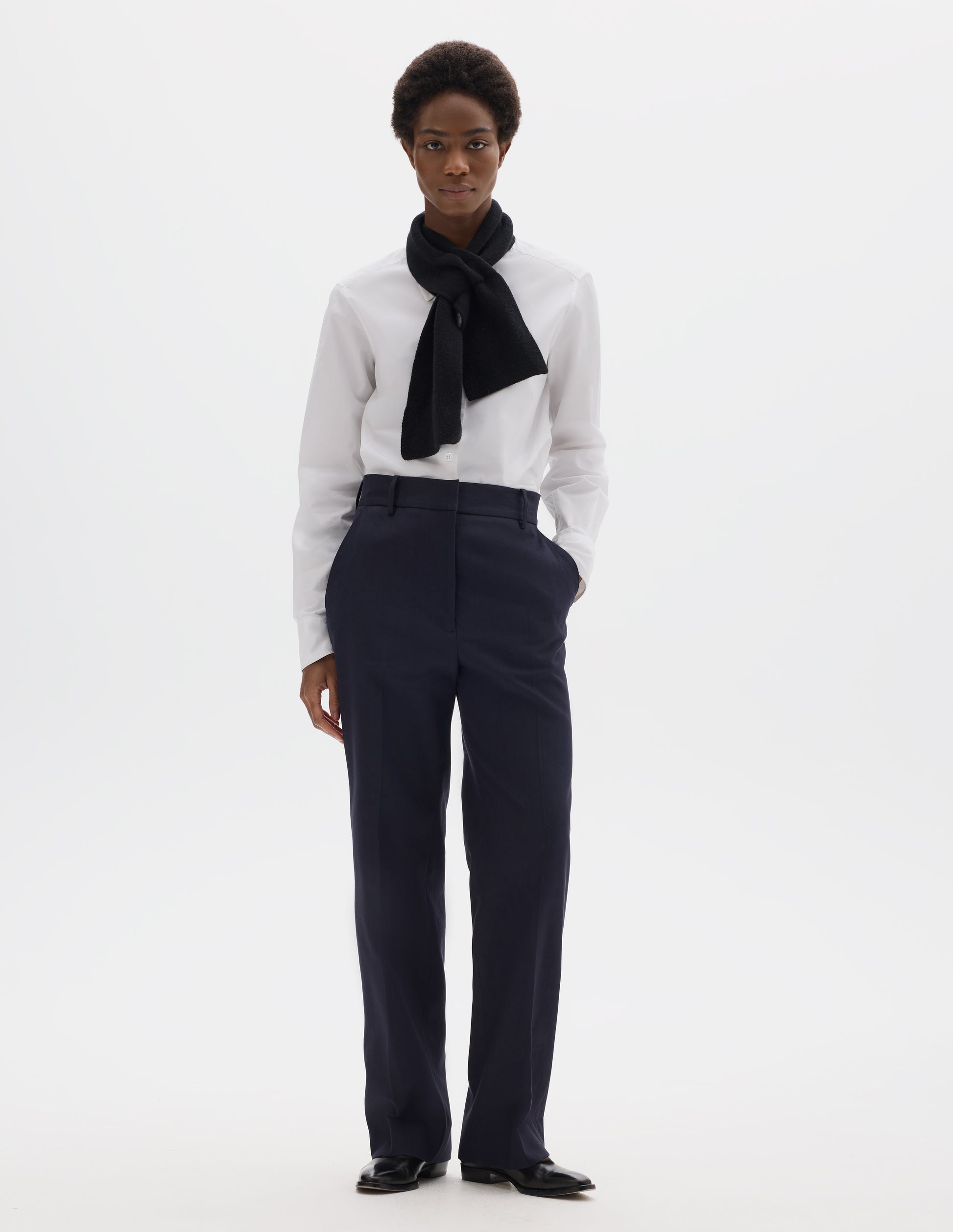 Full body image, front view of model wearing dark blue cotton linen straight leg trousers with belt loops and slant pockets. Styled with white shirt, black scarf and black leather shoes.