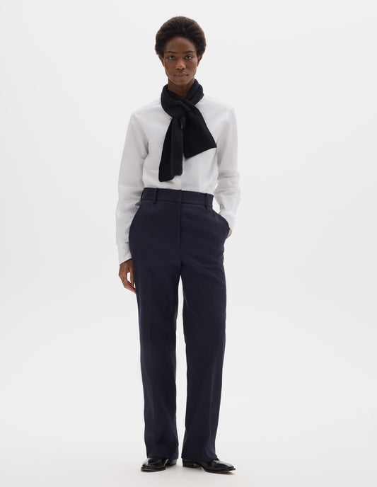 Full body image, front view of model wearing dark blue cotton linen straight leg trousers with belt loops and slant pockets. Styled with white shirt, black scarf and black leather shoes.
