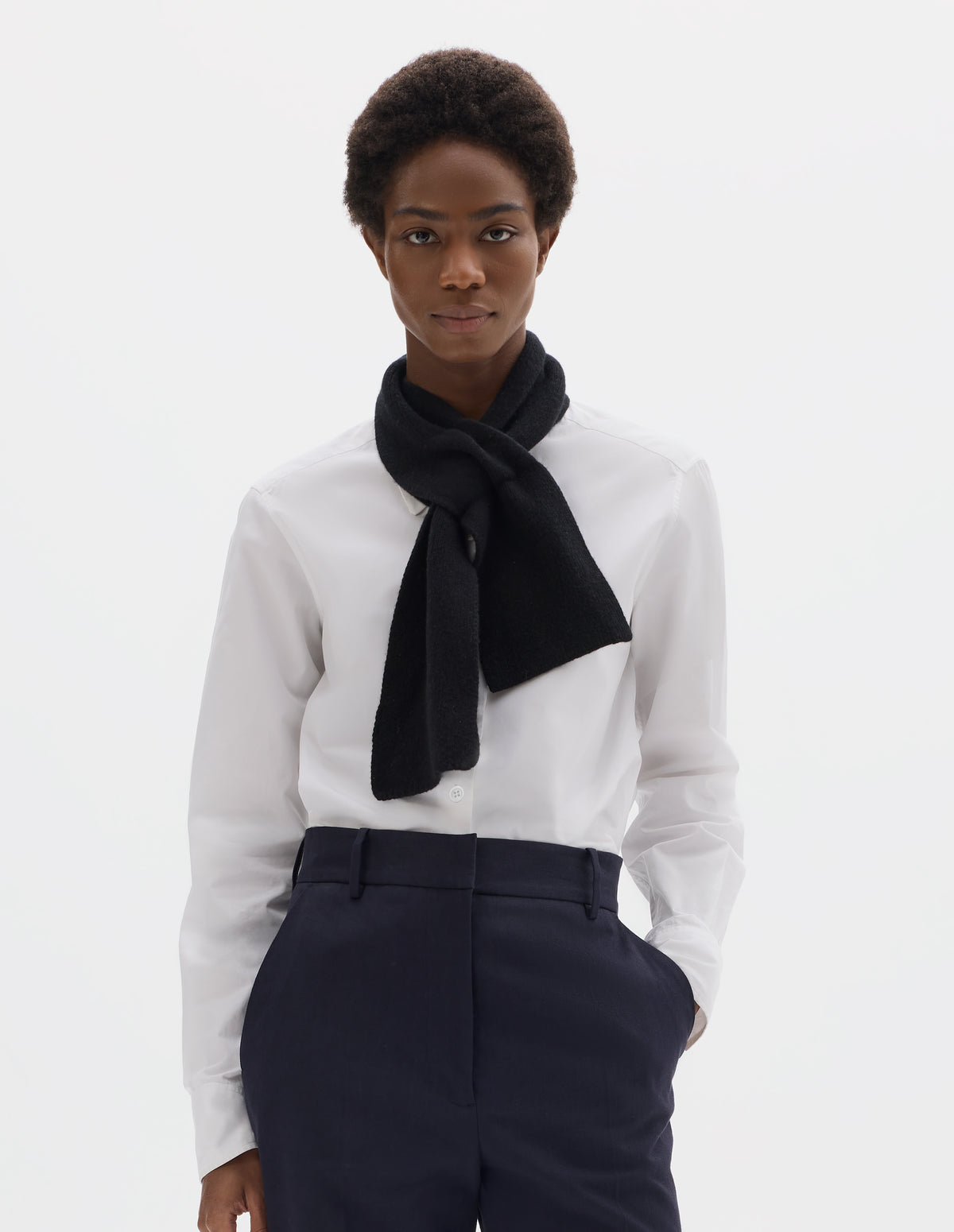 Cropped image, front view of female model wearing black cashmere merino tie through knitted scarf. Styled with white shirt and dark blue trousers.