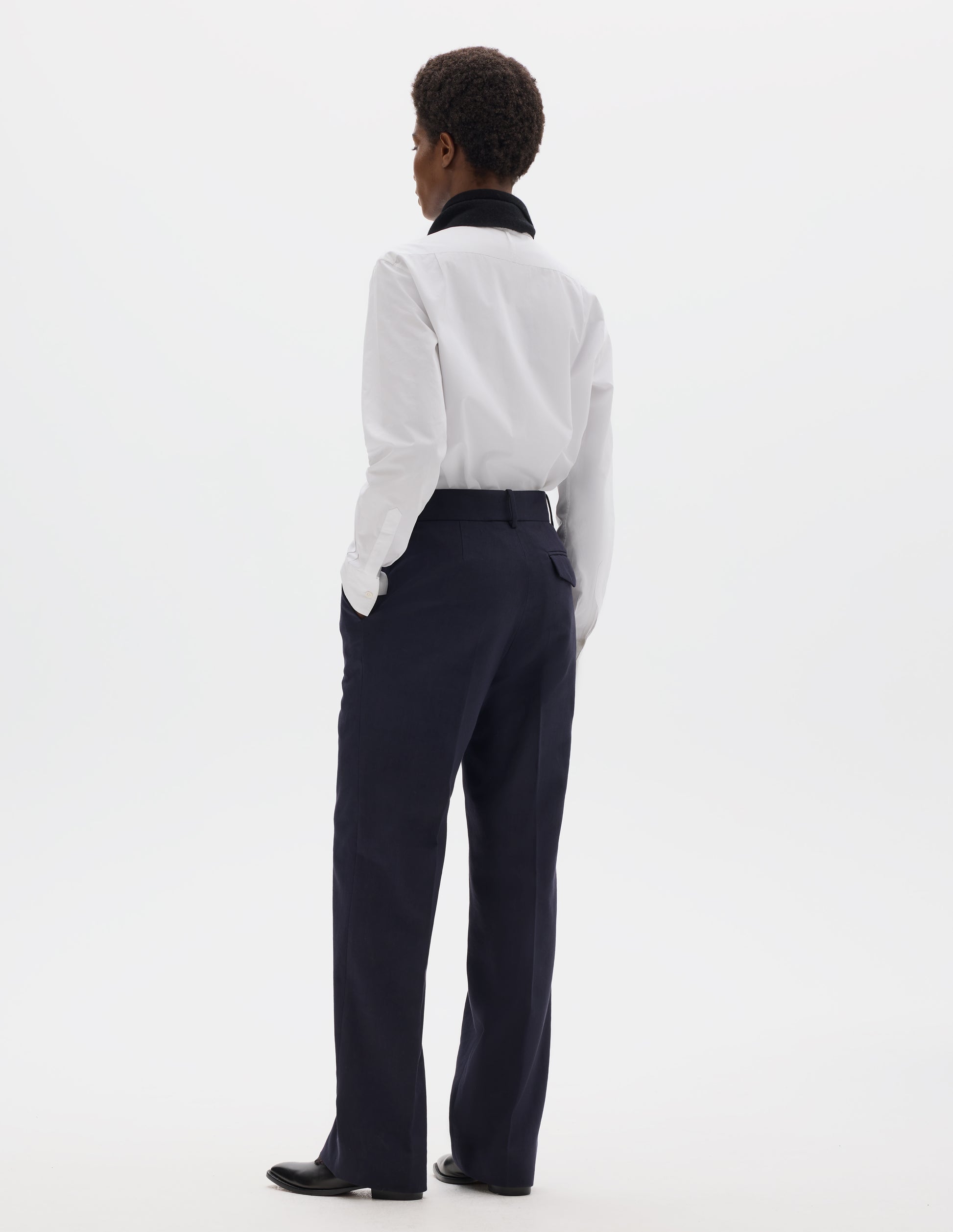 Full body image, back view of model wearing dark blue cotton linen straight leg trousers with belt loops and concealed horn button-fastening flap back pocket. Styled with white shirt, black scarf and black leather shoes.