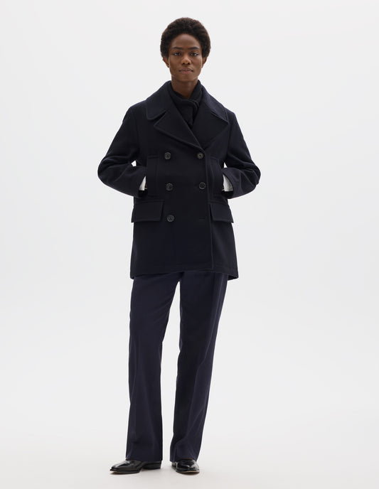 Full body image, front view of model wearing dark blue wool coat with large collar and revere, double breasted front with horn buttons, high-set single welt pockets and large flap pockets. Styled with black knitted scarf, dark blue trousers and black leather shoes.
