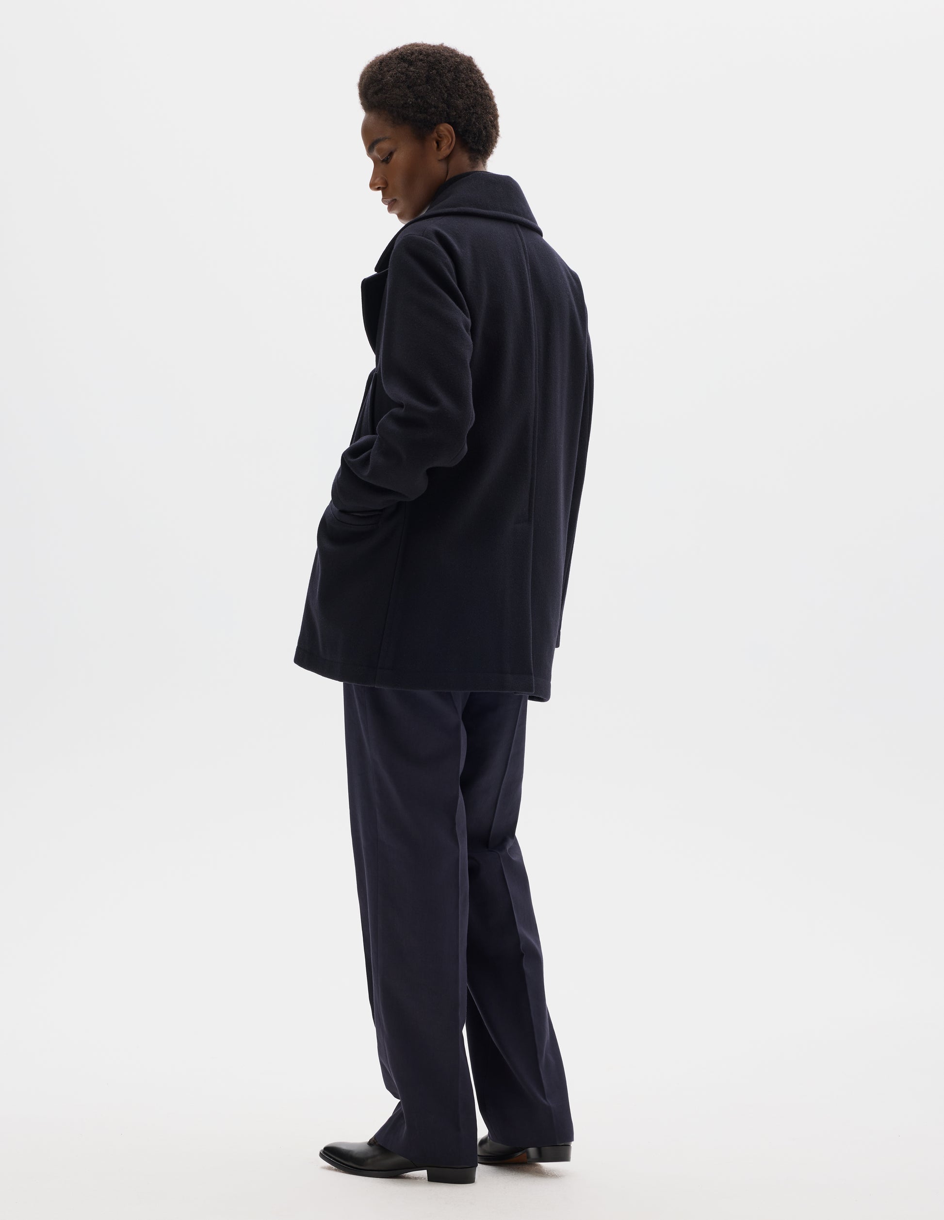 Full body image, back view of model wearing dark blue wool coat with large collar, centre back seam and rear vent. Styled with dark blue trousers and black leather shoes. 