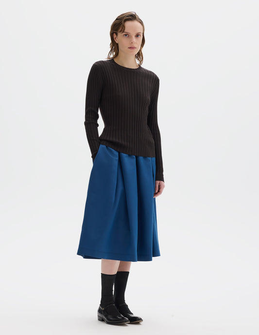 Full body image, front view of model wearing neat fitting dark brown merino top with wide rib through body, wide contrast rib on crew neck, and set-in long sleeves with fine rib cuffs. Styled with blue skirt, black socks and black leather shoes.