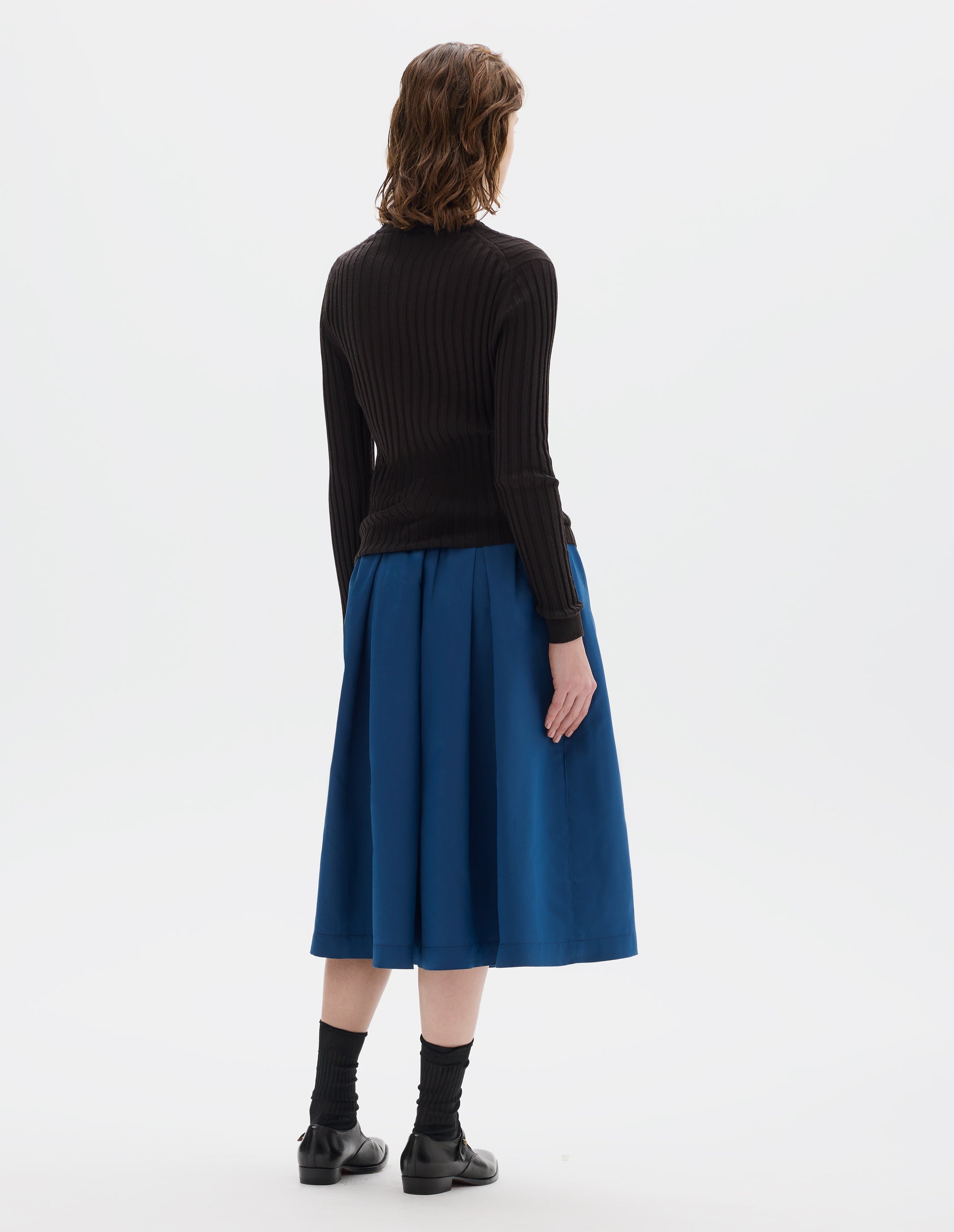 Full body image, back view of model wearing neat fitting dark brown merino top with wide rib through body, wide contrast rib on crew neck, and set-in long sleeves with fine rib cuffs. Styled with blue skirt, black socks and black leather shoes.