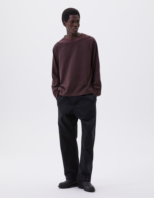 Full body image, front view of model wearing relaxed black trousers with slant welt pockets, corozo brace button fly and press stud hem adjusters. Styled with brown long sleeve t-shirt and black leather boots.
