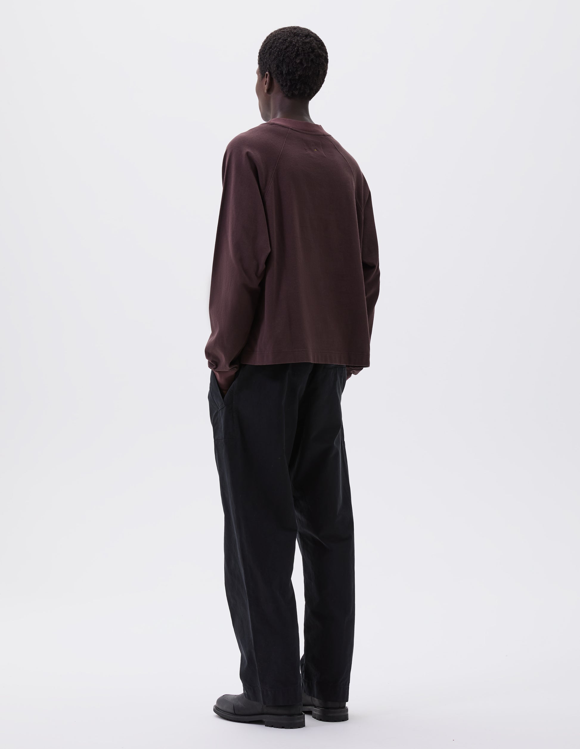 Full body image, back view of model wearing relaxed black trousers with slant welt pockets  and press stud hem adjusters. Styled with brown long sleeve t-shirt and black leather boots. 