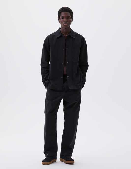 Full body image, front view of model wearing black overshirt with press stud fastenings, stich through chest pocket and adjustable cuffs. Styled with matching black trousers, burgundy t-shirt and black suede t-shirt. 