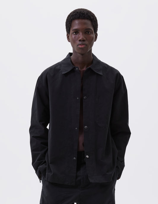 Cropped body image, front view of model wearing black overshirt with press stud fastenings, stich through chest pocket and adjustable cuffs. Styled with matching black trousers and burgundy t-shirt.