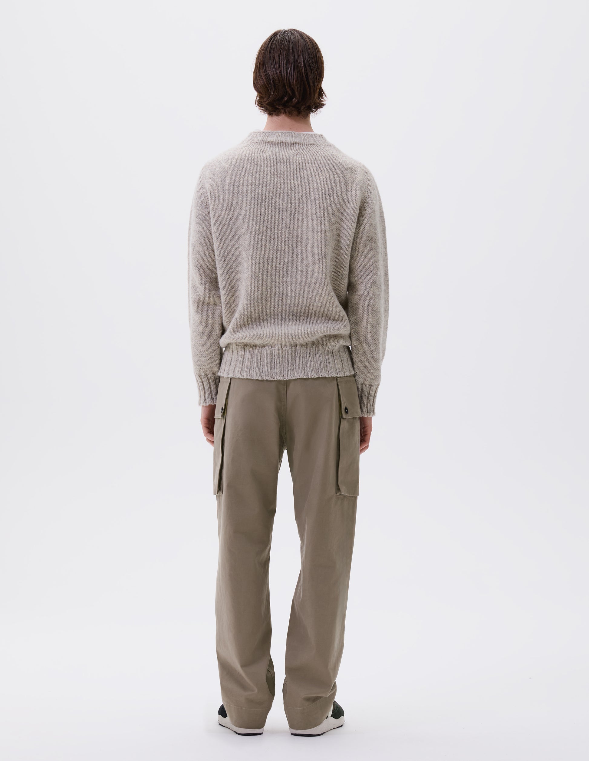 Full body image, back view of model wearing beige cargo trousers with deep flap button fastening patch pocket, belt loops and welt pocket on back yoke. Styled with beige jumper and brown and black trainers.