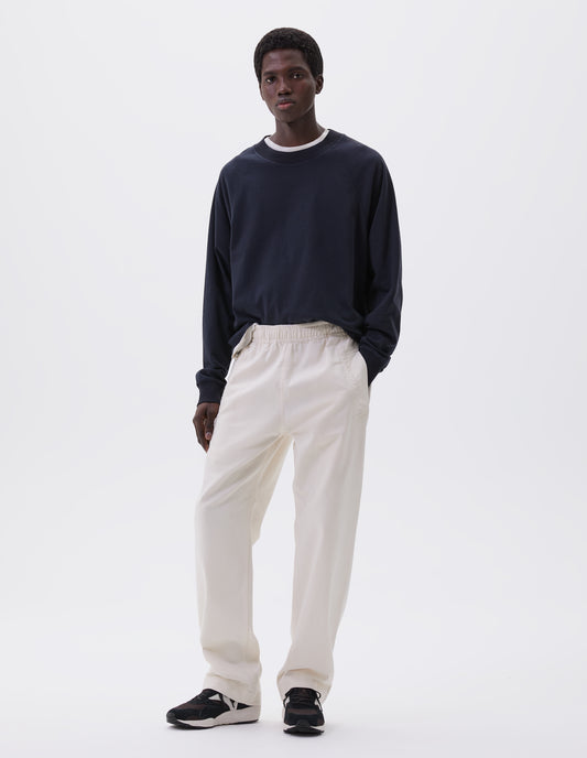 Full body image, front view of model wearing navy blue long sleeve t-shirt with raglan sleeve, single rib trims on neck and cuffs and vent split hem. Styled over a white t-shirt, off white joggers and black and brown trainers.