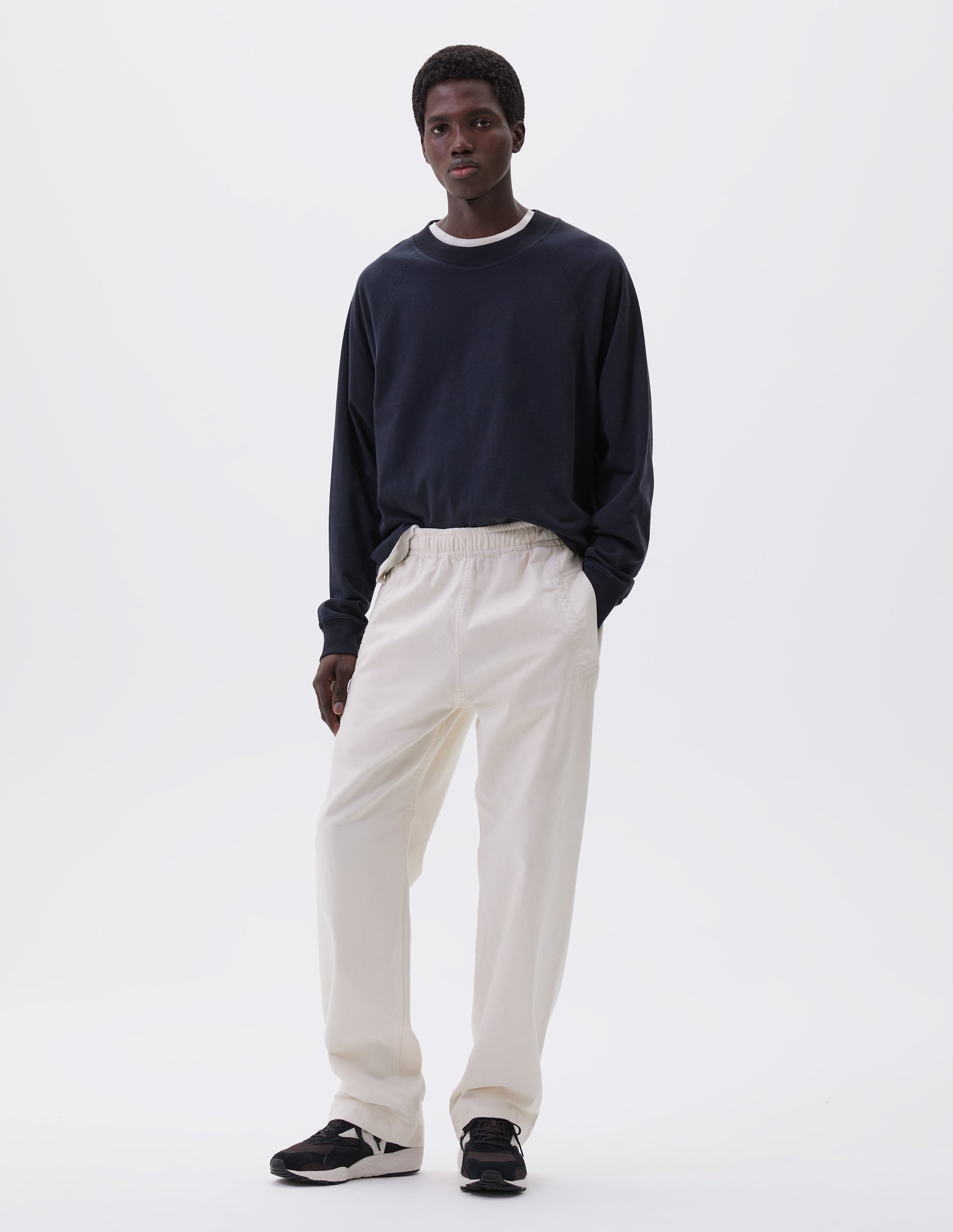 Full body image, front view of model wearing relaxed white joggers with elasticated waist and deep welt pockets. Styled with dark blue long sleeve t-shirt over white t-shirt and with black, brown and white trainers.