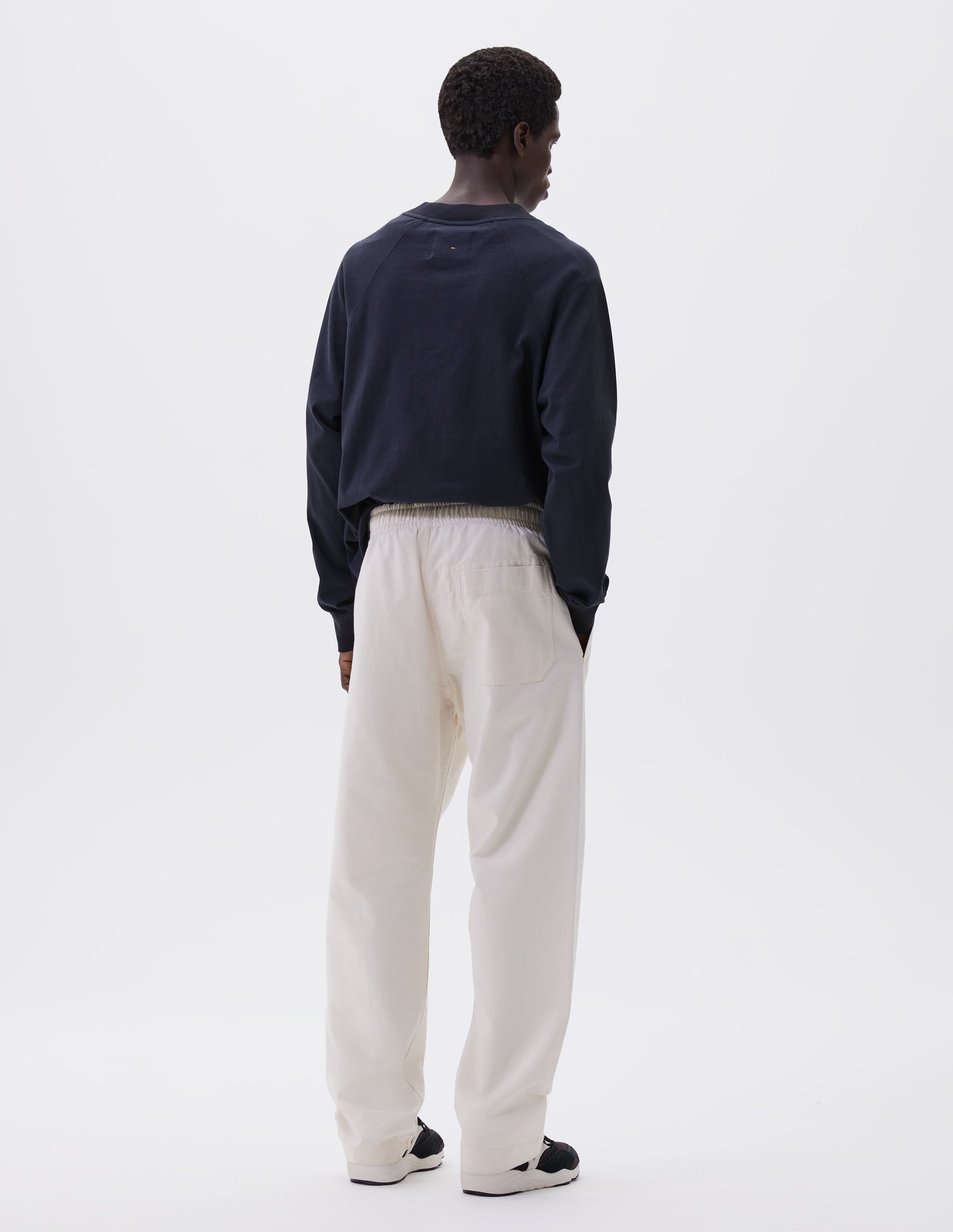Full body image, back view of model wearing relaxed white joggers with elasticated waist and patch back pocket. Styled with dark blue long sleeve t-shirt and black, brown and white trainers. 
