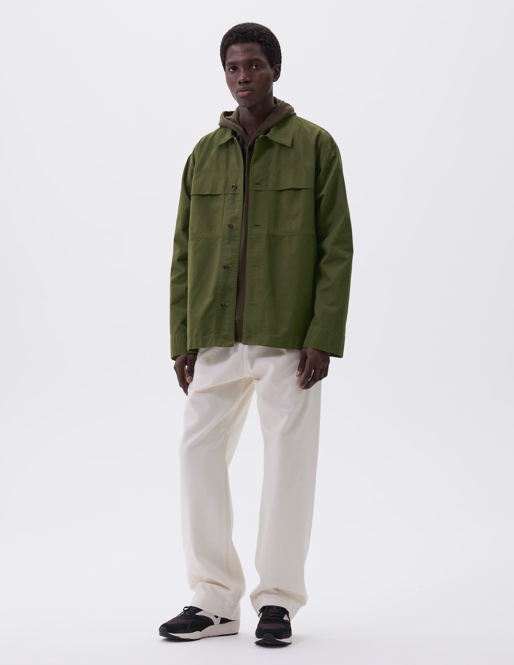 Full body image, front view of model wearing relaxed, overshirt cut green shirt  with large storm flap patch chest pockets, corozo brace buttons, straight cuffs and vent split hem with herringbone tape detailing. Styled over green hoodie, white trousers and black, brown and white trainers.