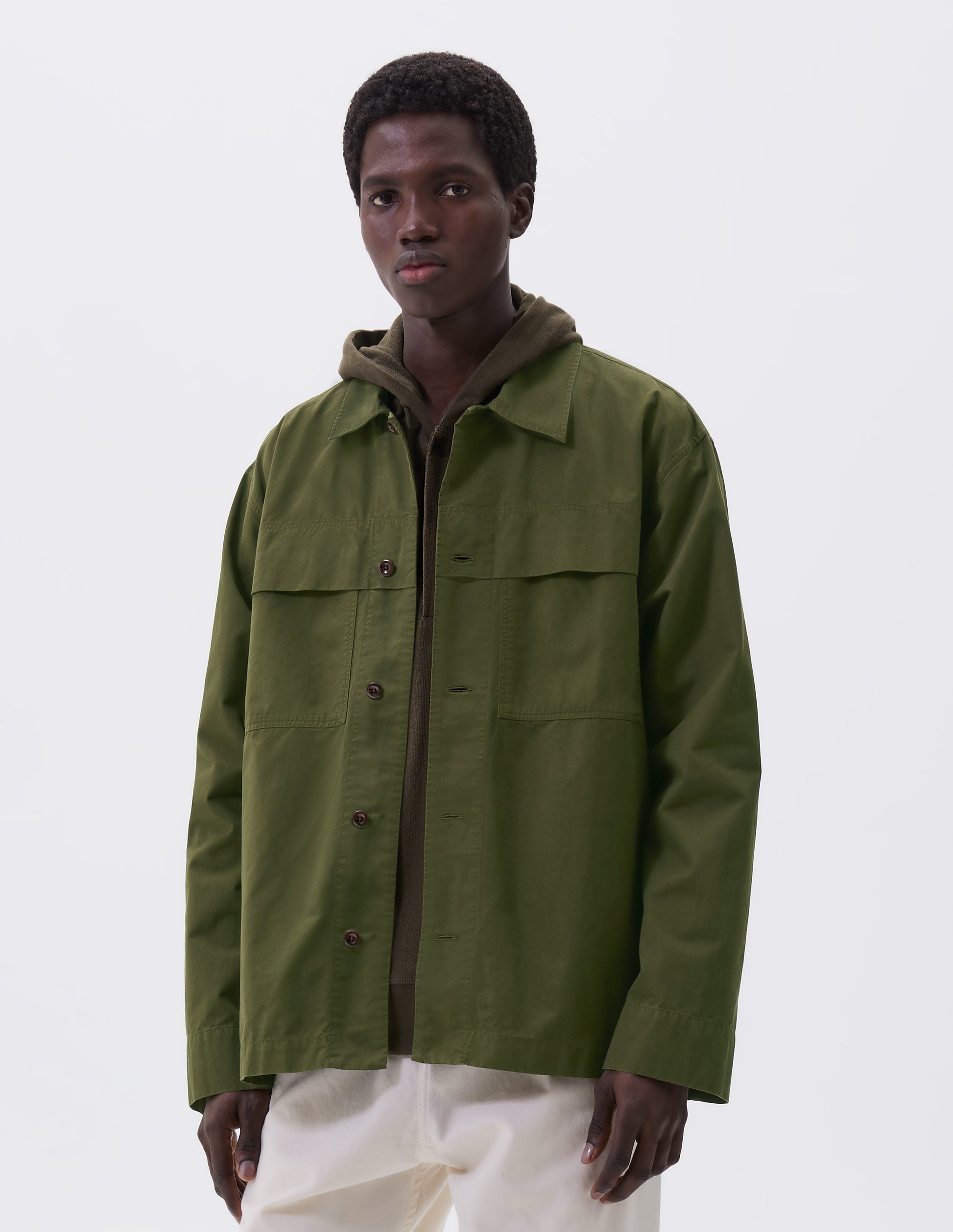 Cropped image, front view of model wearing relaxed overshirt cut green shirt  with large storm flap patch chest pockets, corozo brace buttons, straight cuffs and vent split hem with herringbone tape detailing. Styled over green hoodie and white trousers.