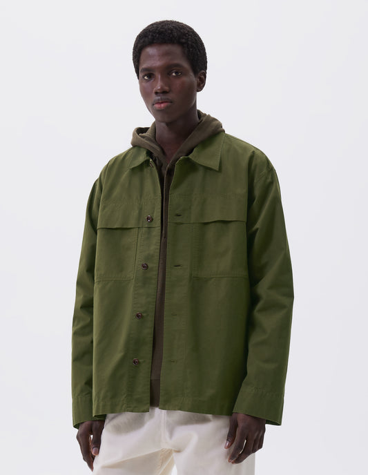Cropped image, front view of model wearing relaxed overshirt cut green shirt  with large storm flap patch chest pockets, corozo brace buttons, straight cuffs and vent split hem with herringbone tape detailing. Styled over green hoodie and white trousers.