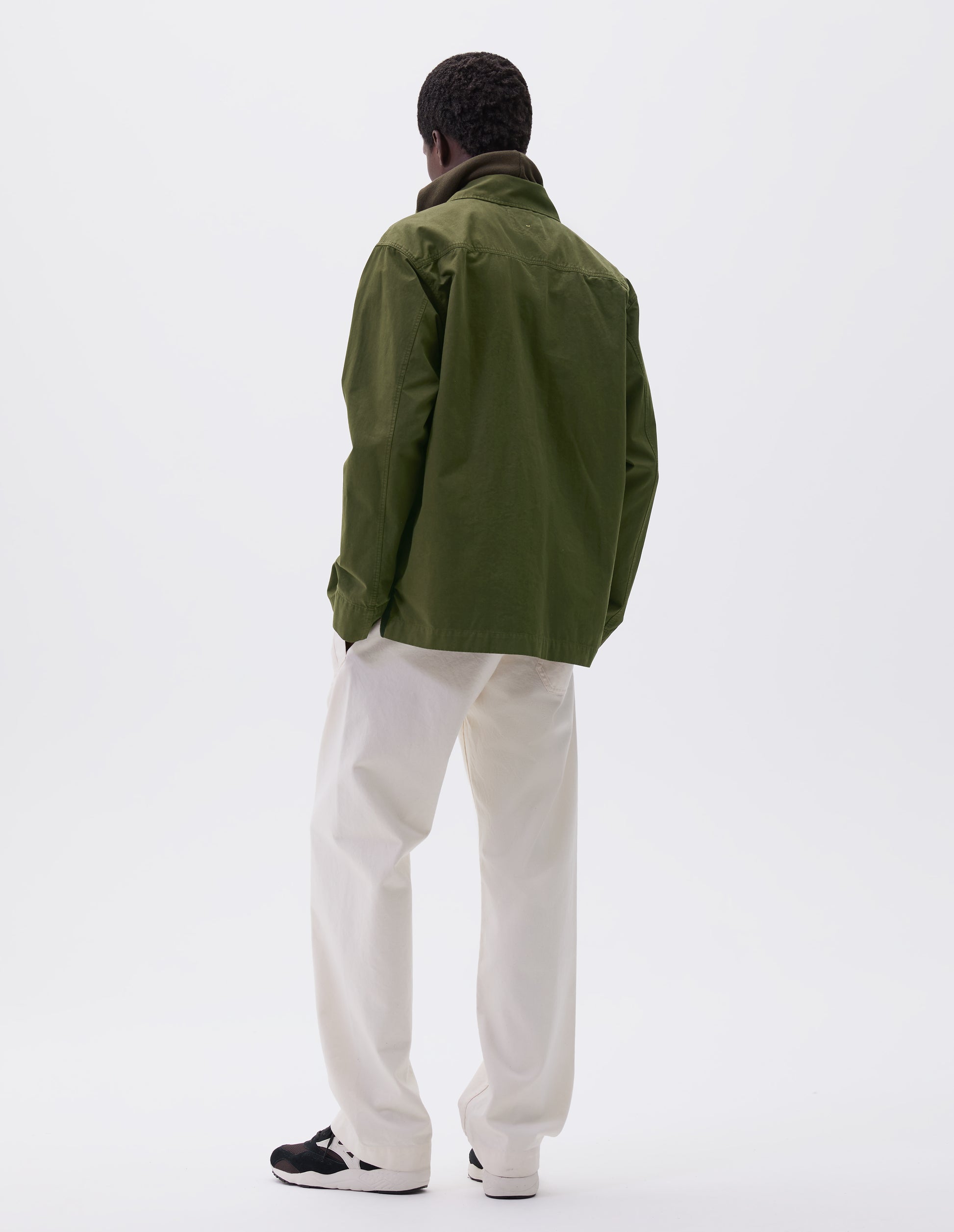 Full body image, back view of model wearing relaxed, overshirt cut green shirt with straight cuffs and vent split hem with herringbone tape detailing. Styled over green hoodie, white trousers and black, brown and white trainers.