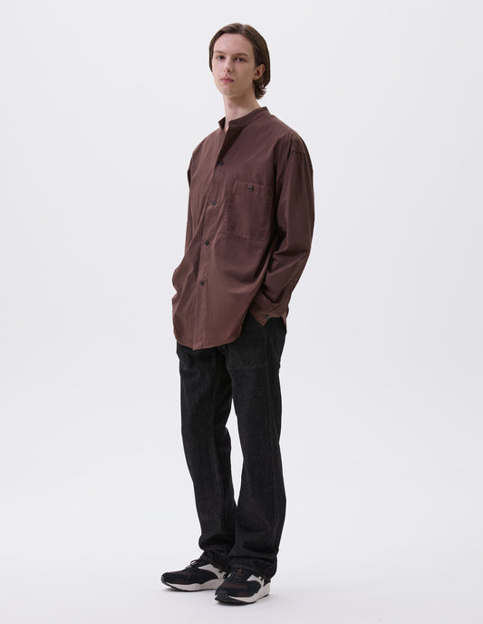Full body image, front view of model wearing brownish red cotton collarless shirt, with stitch through placket, button-through patch pocket and curved side gusset. Styled with black denim jeans and brown and black trainers.