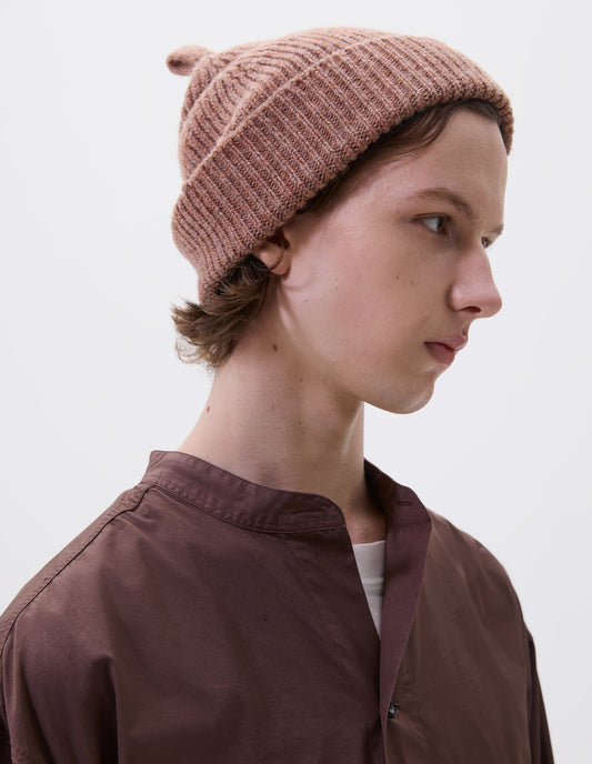 Cropped body image, side view of male model wearing pinkish beige wool ribbed hat, with knitted loop detailing on top. Styled with brownish red collarless shirt.