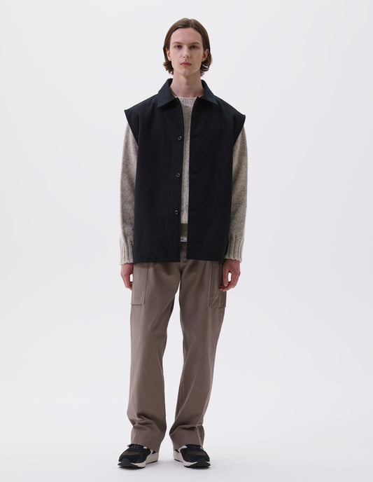 Full body image, front view of model wearing black sleeveless wool cotton drill overshirt with stitch through facing and vent split hem. Styled with light beige jumper, dark beige trousers and brown and black trainers.