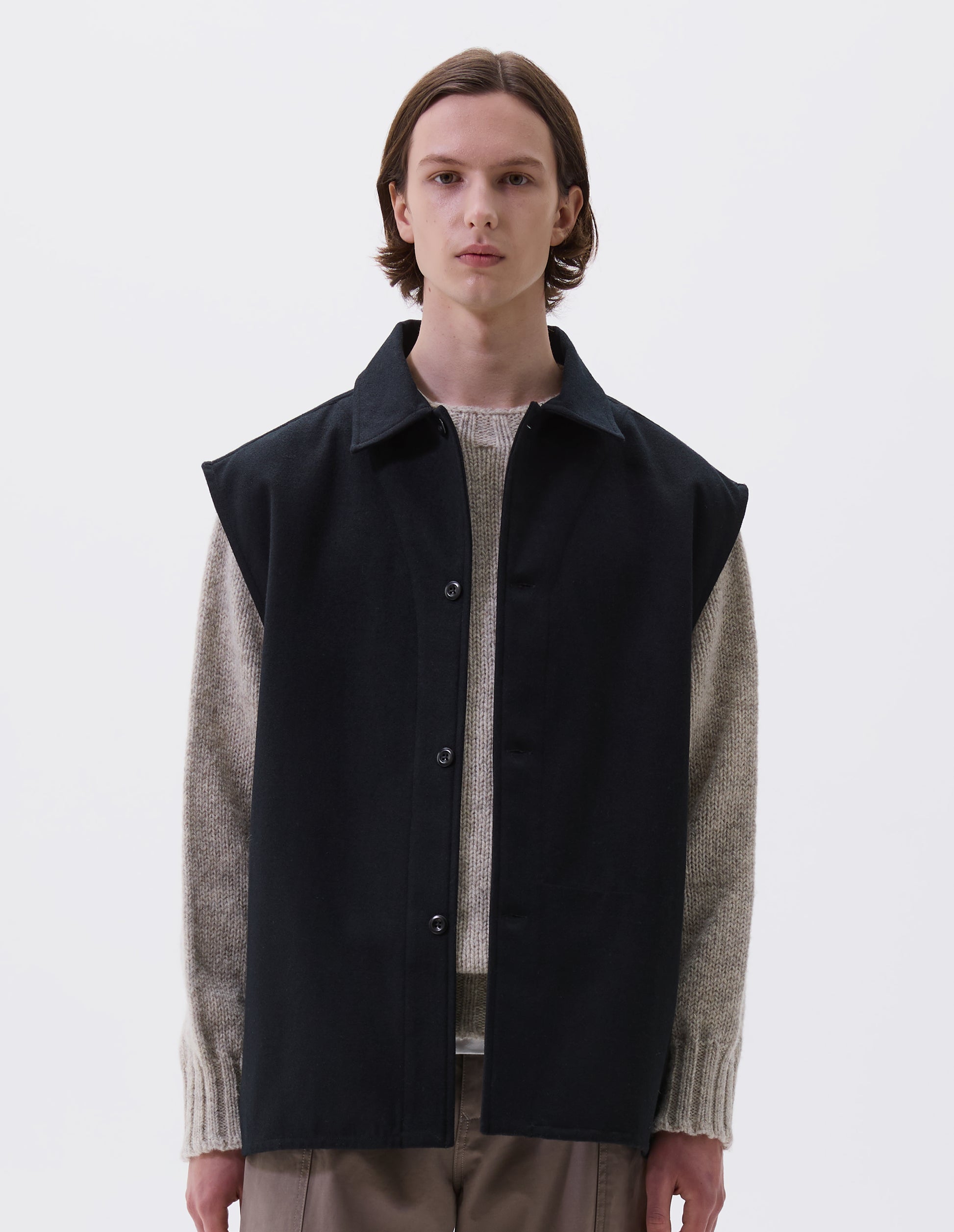 Cropped body image, front view of model wearing black sleeveless wool cotton drill overshirt with stitch through facing and vent split hem. Styled with light beige jumper and dark beige trousers.