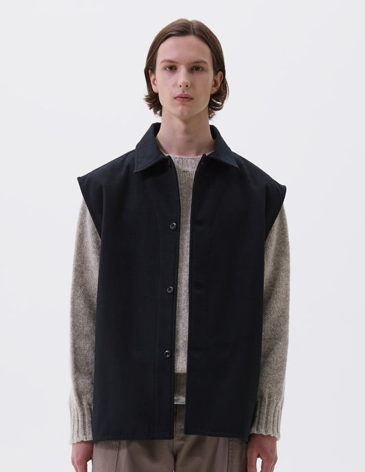 Cropped body image, front view of model wearing black sleeveless wool cotton drill overshirt with stitch through facing and vent split hem. Styled with light beige jumper and dark beige trousers.