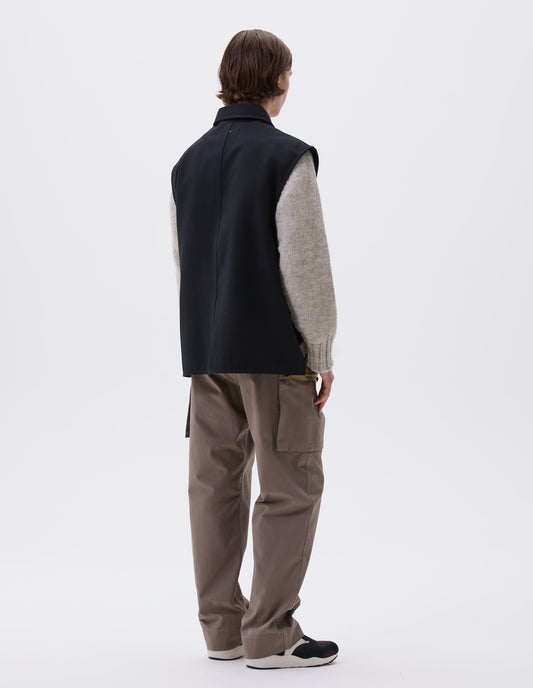 Full body image, back view of model wearing black sleeveless wool cotton drill overshirt with stitch through facing and vent split hem. Styled with light beige jumper, dark beige trousers and brown and black trainers.