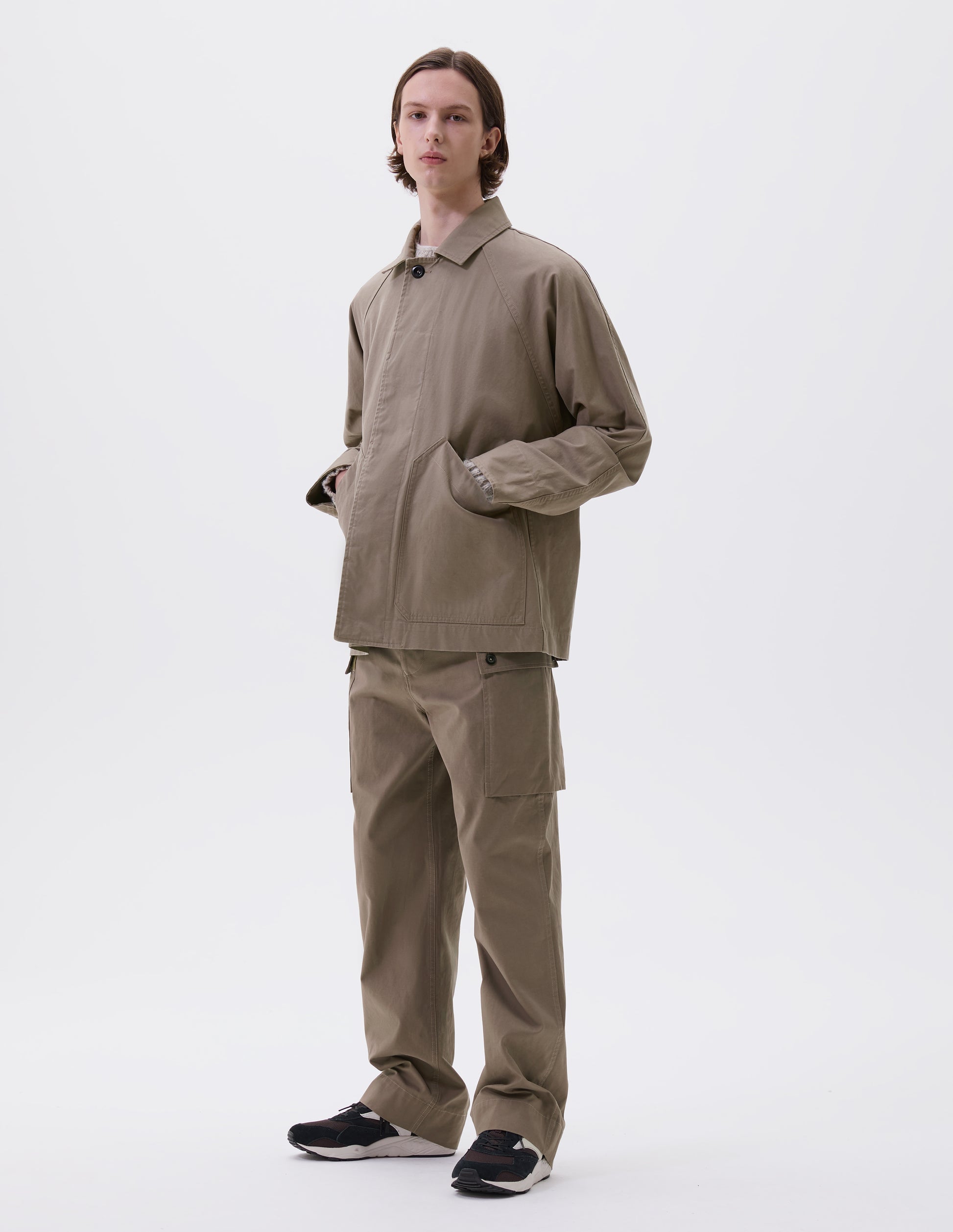 Full body image, front view of model wearing beige raglan sleeve jacket with corozo button fastening fly front and slant patch pockets. Styled over a chunky beige jumper, with matching trousers and black and brown trainers.