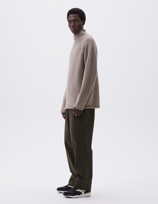 Full body image, front view of model wearing green brown trousers with button fly, deep stitch hem and exposed double rivet. Styled with chunky high neck beige jumper and brown and black trainers.