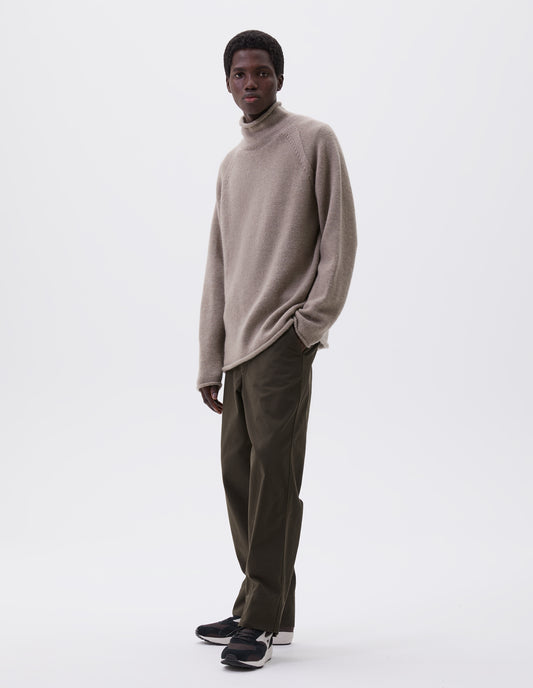 Full body image, front view of model wearing beige roll neck jumper with seamless construction, high neck, raglan sleeve and straight cuffs and hem. Styled with green trousers and black and brown trainers.