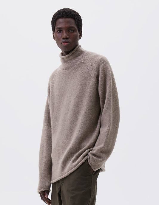 Cropped body image, front view of model wearing beige roll neck jumper with seamless construction, high neck, raglan sleeve and straight cuffs and hem. Styled with green trousers.