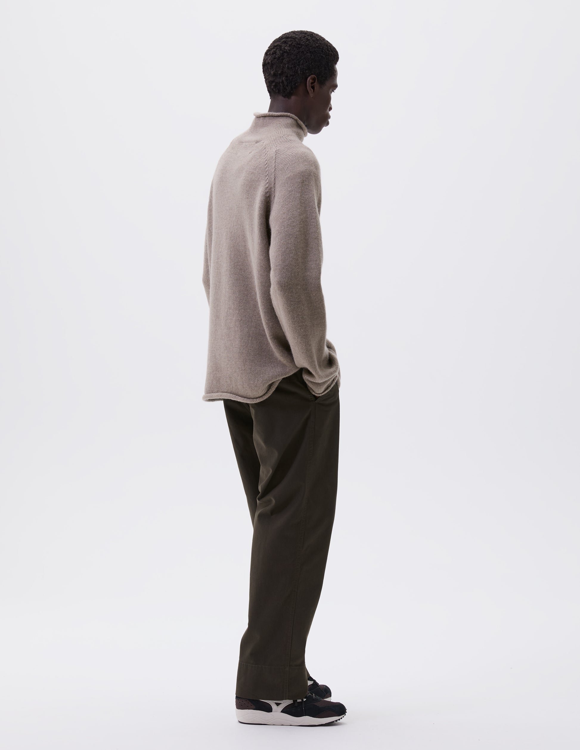 Full body image, back view of model wearing beige roll neck jumper with seamless construction, high neck, raglan sleeve and straight cuffs and hem. Styled with green trousers and black and brown trainers.