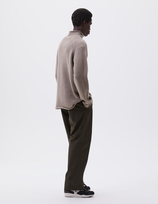 Full body image, back view of model wearing beige roll neck jumper with seamless construction, high neck, raglan sleeve and straight cuffs and hem. Styled with green trousers and black and brown trainers.