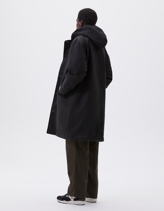 Full body image, back view of model wearing black hooded parka with fleece lined drawcord hood, two way zip fastening and slanted flap pockets. Styled with green trousers and black and brown trainers. 