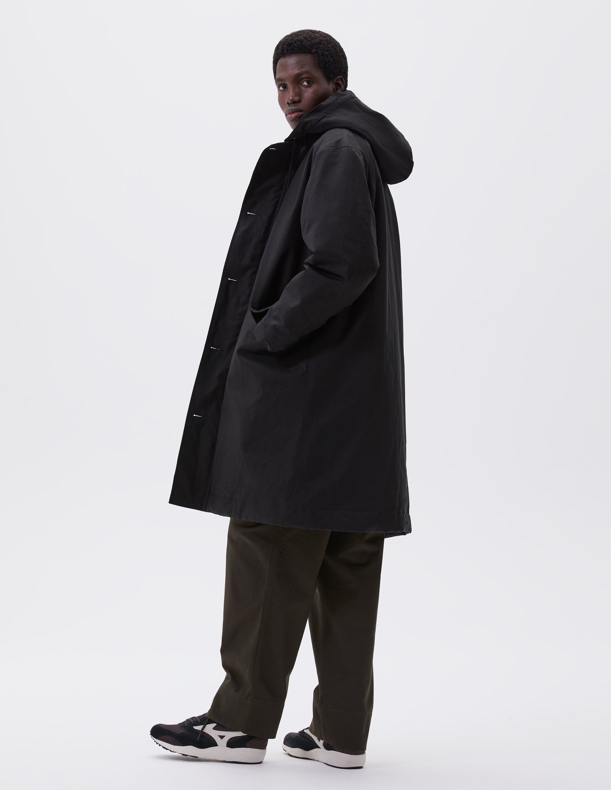 Full body image, side view of model wearing black hooded parka with fleece lined drawcord hood, two way zip fastening and slanted flap pockets. Styled with green trousers and black and brown trainers. 