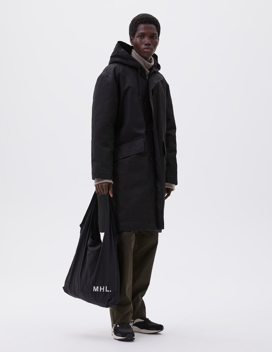 Full body image, front view of model wearing black hooded parka with fleece lined drawcord hood, two way zip fastening with button fastening storm flap and slanted flap pockets. Styled with beige high neck jumper, green trousers, MHL black branded tote bag and black and brown trainers. 