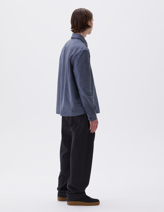 Full body image, back view of model wearing blue long sleeve shirt, with corozo button cuffs. Styled with indigo blue joggers and black nubuck leather trainers.