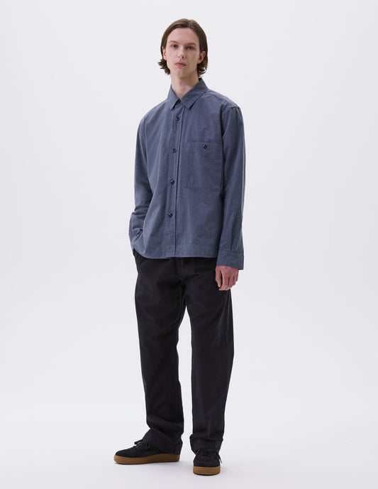 Full body image, front view of model wearing blue long sleeve shirt, with raised placket, corozo button closure and chest patch pocket. Styled with indigo blue joggers and black nubuck leather trainers.