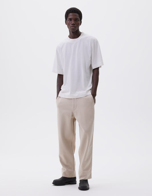 Full body image, front view of model wearing off white t-shirt, with rib neck and flatlock stitch detailing on seams. Styled with off white trousers and black leather boots.