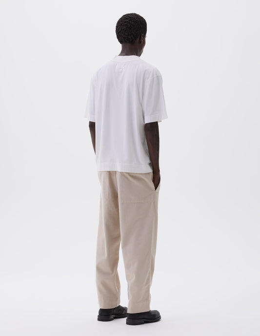 Full body image, back view of model wearing off white t-shirt, with rib neck and flatlock stitch detailing on seams. Styled with off white trousers and black leather boots.