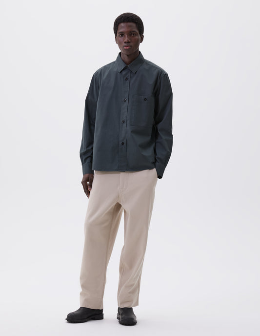 Full body image, front view of model wearing blue green long sleeve shirt, with raised placket, button through chest patch pocket and corozo brace button closure and cuffs. Styled with off white trousers and black leather boots