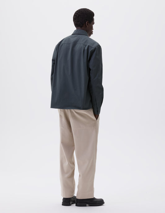 Full body image, back view of model wearing blue green long sleeve shirt, with corozo brace button cuffs. Styled with off white trousers and black leather boots.