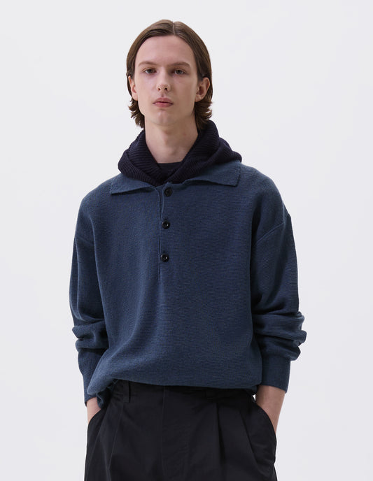 Cropped body image, front view of model wearing dark navy blue wool hood with contrasting black herringbone tape to adjust and contrast rib on face. Styled with blue knitted polo and black pleated trousers.