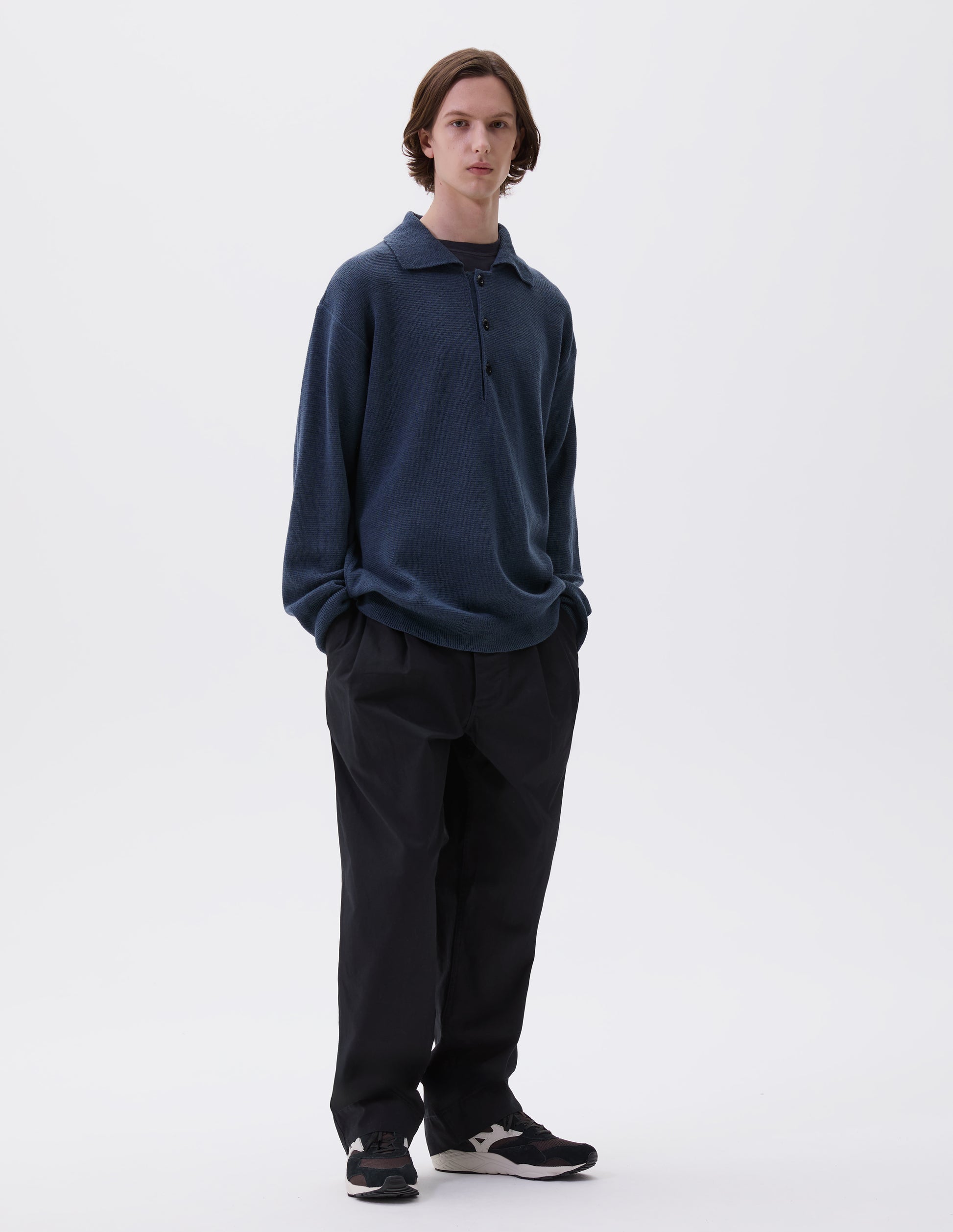 Full body image, front view of model wearing blue knitted polo with contrast rib on neck, cuffs and hem and corozo brace buttons. Styled over navy blue t-shirt, with black trousers and black and brown trainers.