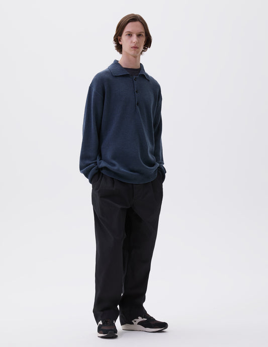 Full body image, front view of model wearing blue knitted polo with contrast rib on neck, cuffs and hem and corozo brace buttons. Styled over navy blue t-shirt, with black trousers and black and brown trainers.