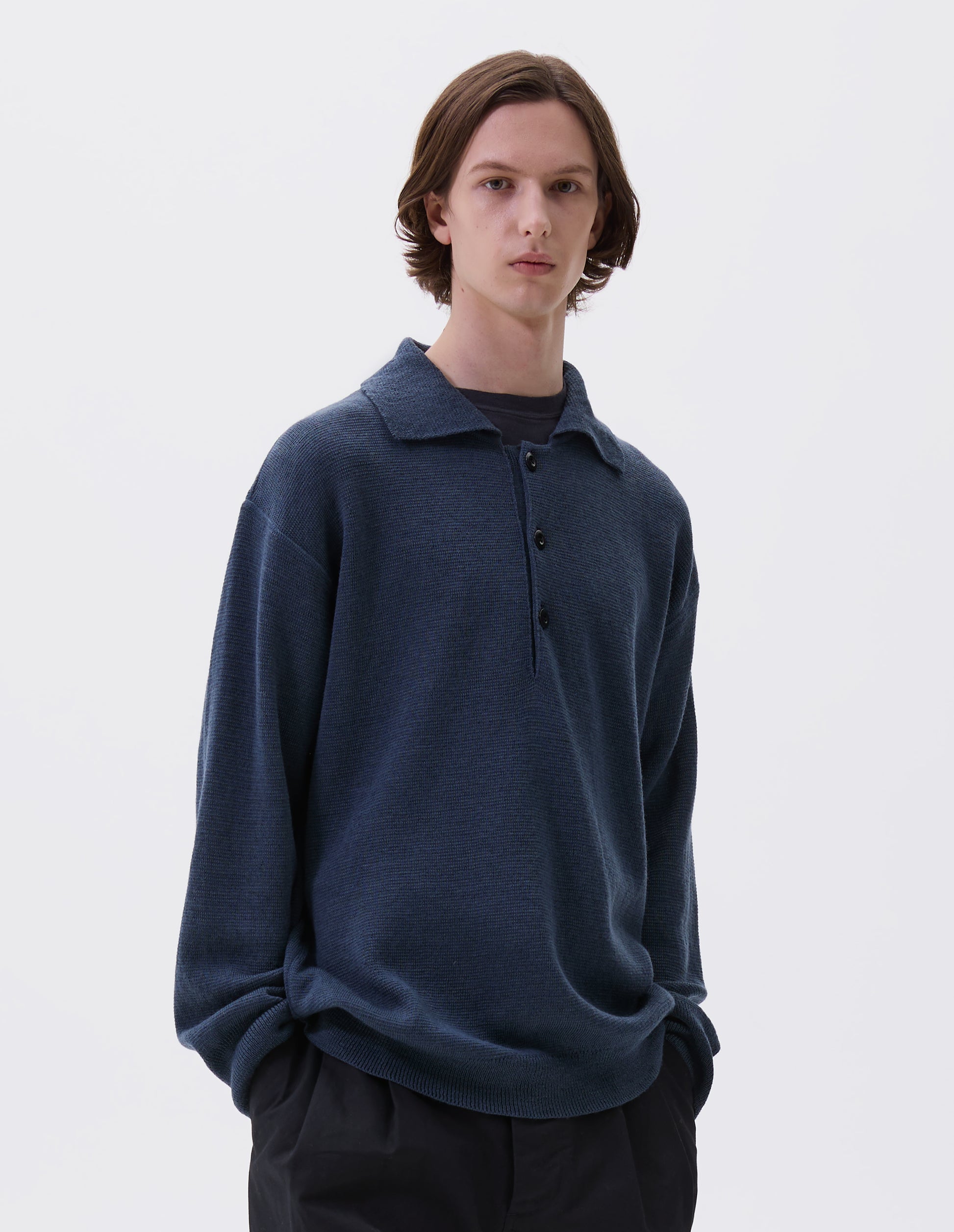 Cropped body image, front view of model wearing blue knitted polo with contrast rib on neck, cuffs and hem and corozo brace buttons. Styled over navy blue t-shirt, with black trousers.