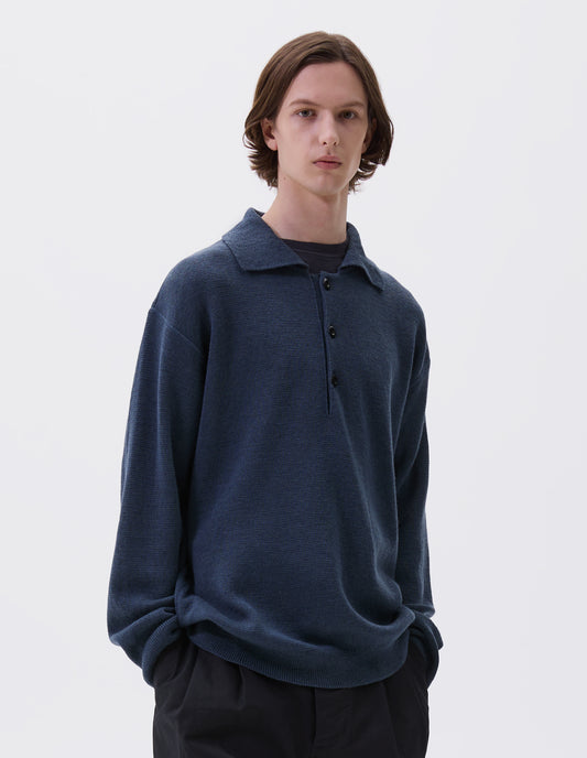 Cropped body image, front view of model wearing blue knitted polo with contrast rib on neck, cuffs and hem and corozo brace buttons. Styled over navy blue t-shirt, with black trousers.