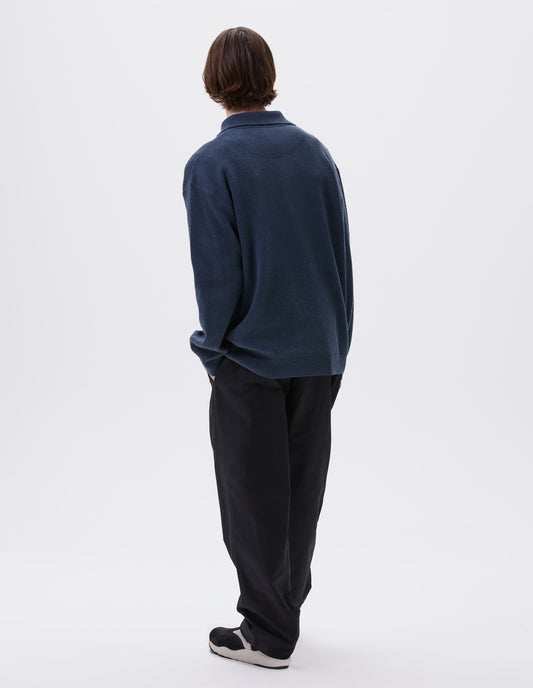 Full body image, back view of model wearing blue knitted polo with contrast rib on neck, cuffs and hem. Styled with black trousers and black and brown trainers.